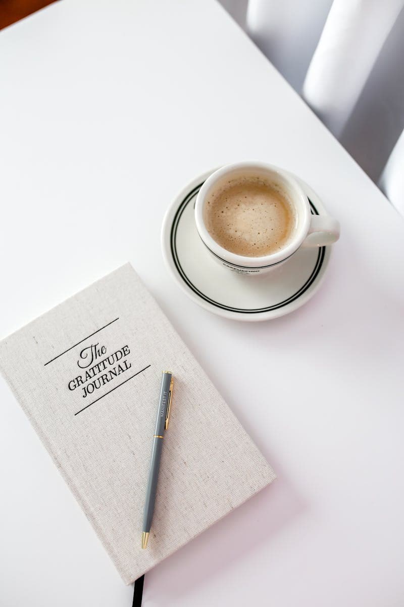 A gratitude journal beside a cup of coffee on a bright, minimalist table setting.